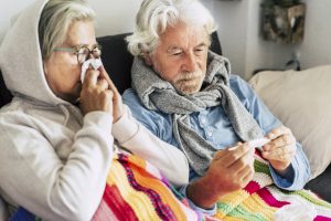 Picture of an older couple sitting under blankets on a couch. The woman is blowing her nose and the man is checking a thermometer.