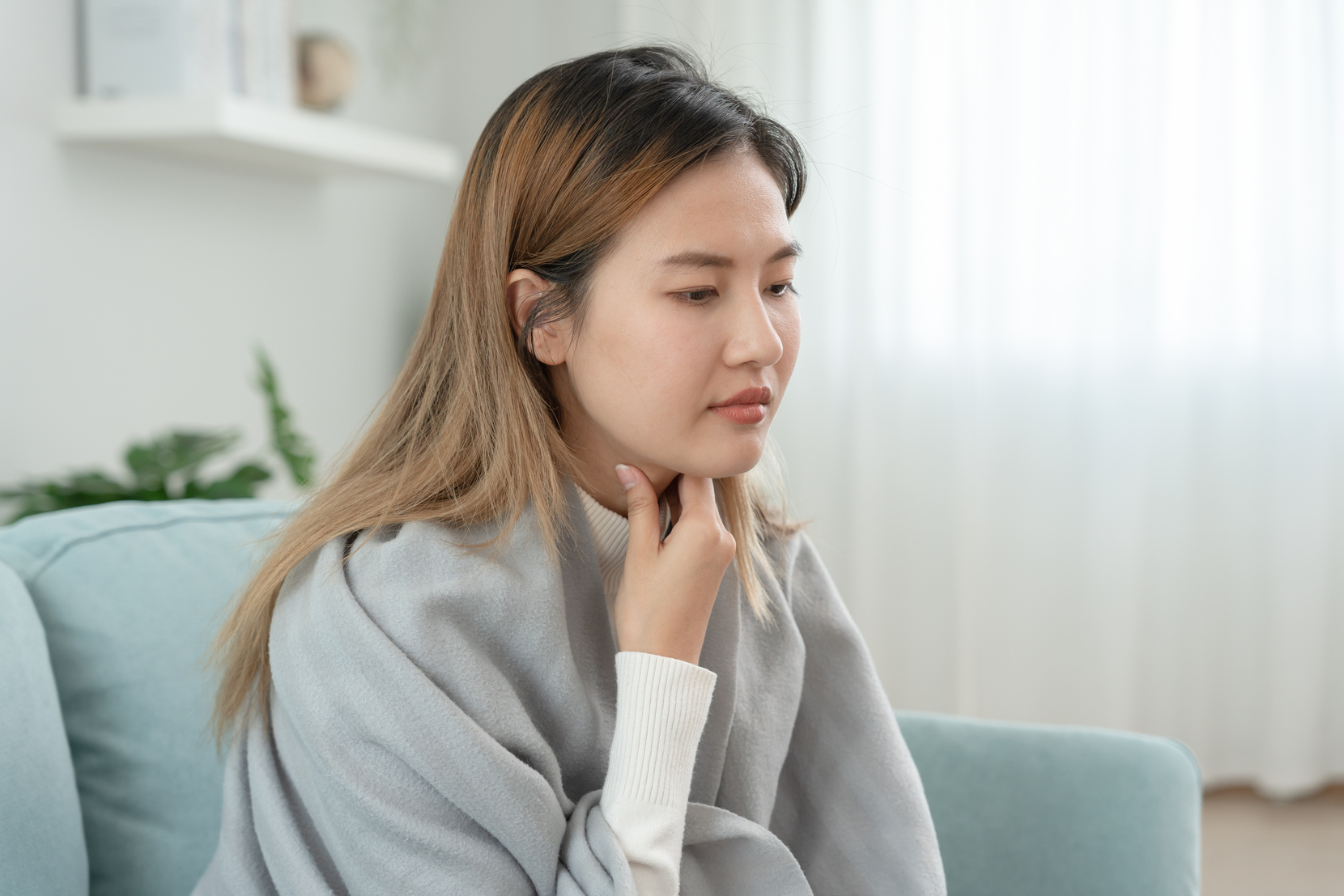 Picture of a woman with a sore throat sitting on a couch and touching her neck in pain.