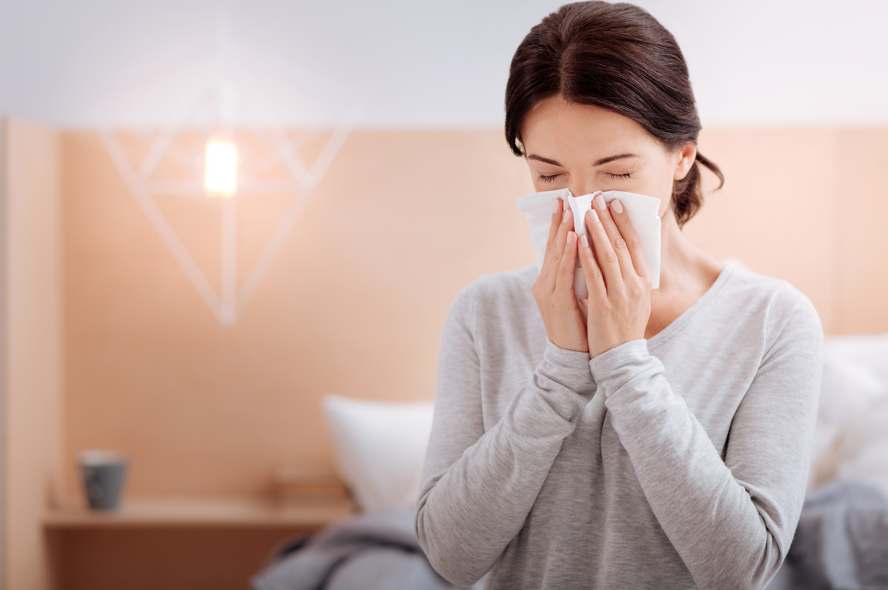 Picture of a sick woman sitting on a bed and sneezing into a tissue.