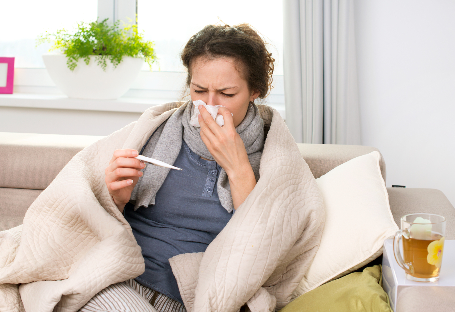 Picture of a woman sneezing into a tissue and looking at a thermometer while sitting under a blanket on a couch.