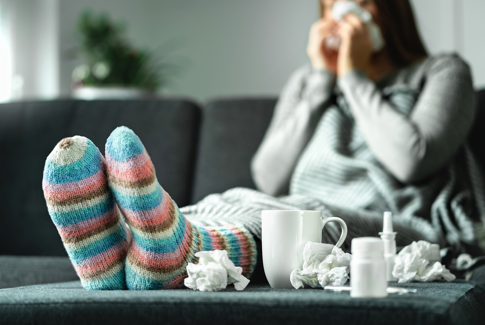 Woman sits on the couch surrounded by tissues, medicine, and a mug as she deals with the flu.