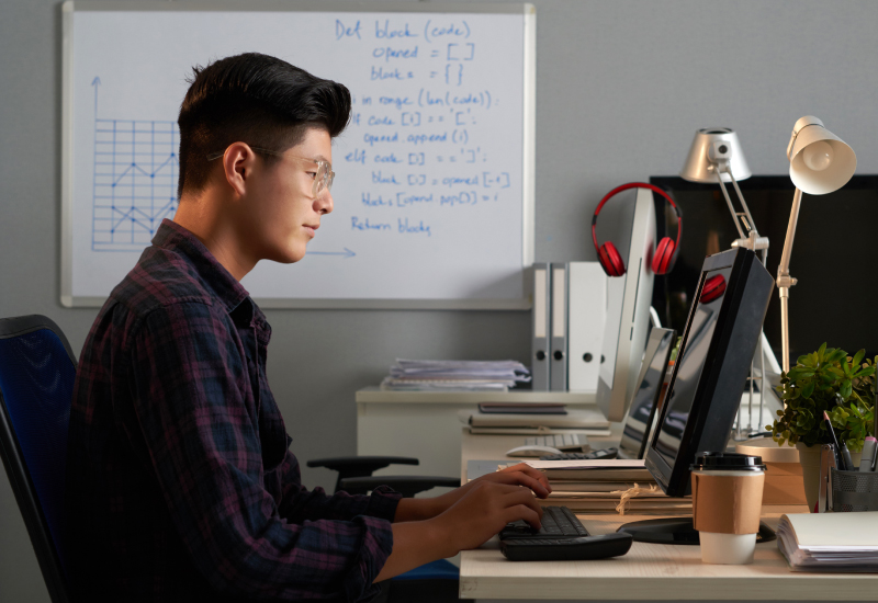 A male freelancer working on his desktop computer at a home office with a whiteboard hanging on a wall on his left side. His work desk has a lamp, coffee in a takeaway cup, some files and a pair of red headphones hanging nearby.