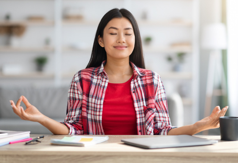 A young woman wearing an unbuttoned checkered shirt over a red t-shirt sitting at her work desk, closing her eyes as if meditating.