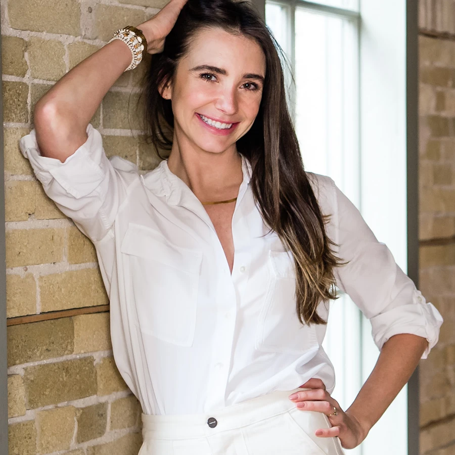 Photo of Blaine in a white outfit in a loft-like building, standing, smiling, touching her hair.