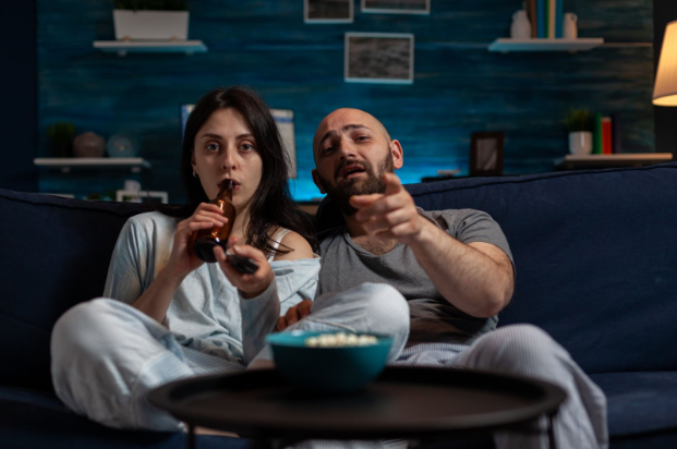 Man and woman sitting on couch, watching TV. Man is pointing at the TV while woman is drinking a beverage and pointing the remote at the TV