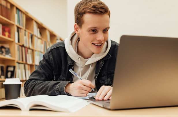 A young man smiling looking at his laptop while sitting in a library taking notes. 