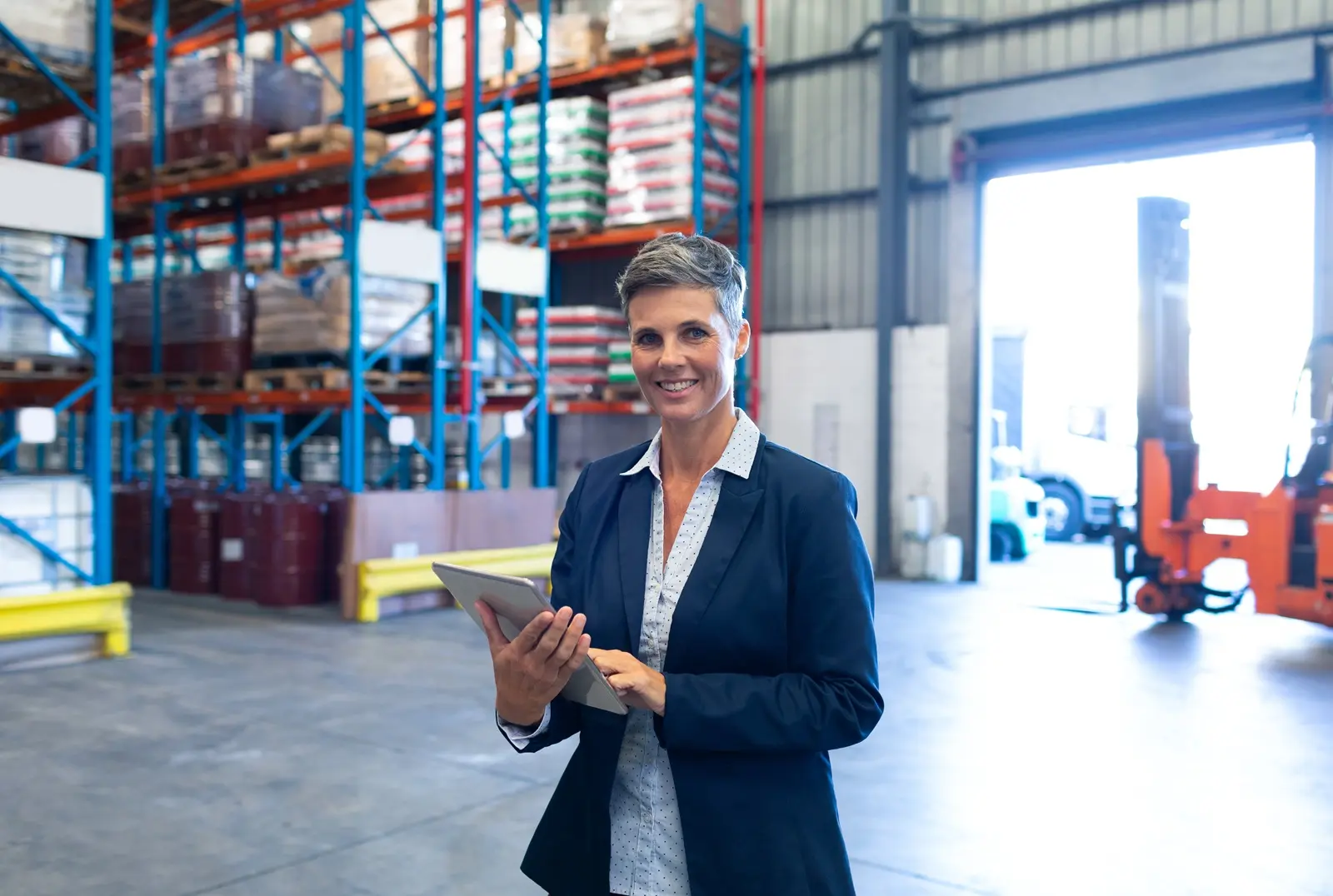 A woman in a blue suit jacket holding a tablet, standing in an active warehouse with a forklift and pallets of goods in the background