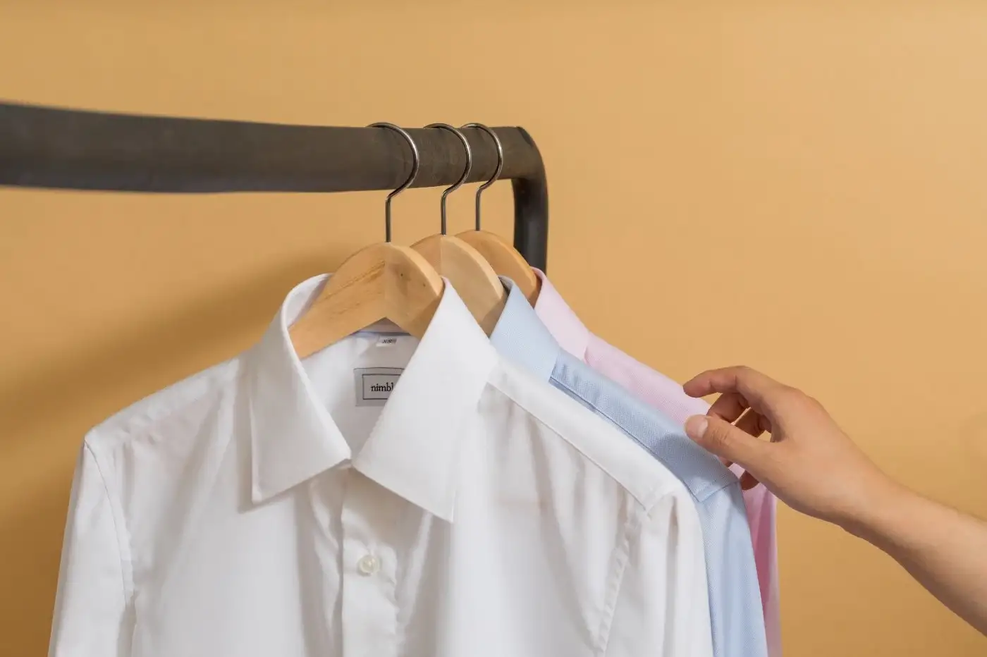 A hand reaching for a blue shirt hung on a clothing rack in between white and pink shirts.