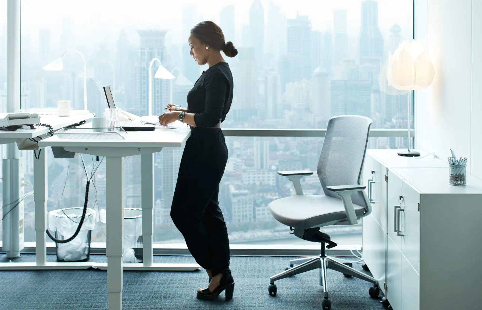 Femme debout à un bureau et prenant des notes devant une grande fenêtre donnant sur une ville.