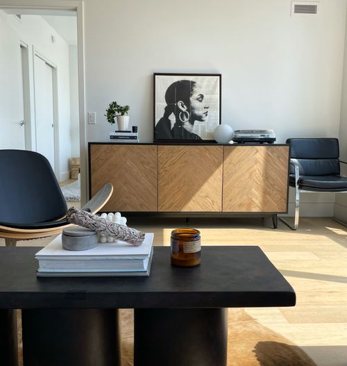 This modern living room features an eye-catching portrait of a powerful Indigenous woman that adds warmth and depth to the space. A neutral colour palette of greys, whites and mauves is used throughout, combined with an abundance of textures such as velvet upholstery and quilted throws. Desktop plants provide a splash of life and greenery while a natural log accent brings in rustic elements. The portrait serves as an unexpected element that is both meaningful and striking. An oversized sectional wrapped in luxurious white fabric enhances the lightness of the room, creating a comfortable yet sophisticated atmosphere. This modern space is both inviting and inspiring for its occupants.