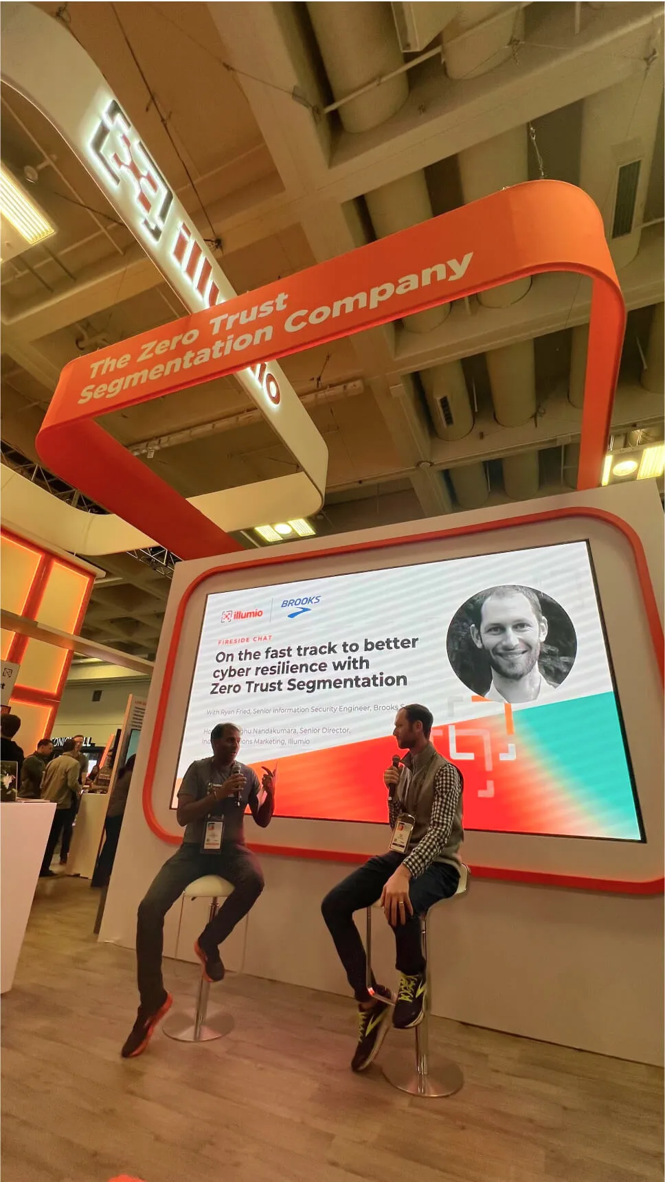 Two men at a cybersecurity convention having a discussion in front of an Illumio booth that reads 'On the fast track to better cyber resilience with zero trust segmentation.'
