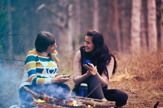 Two women sitting in front of the fire outdoors having coffee