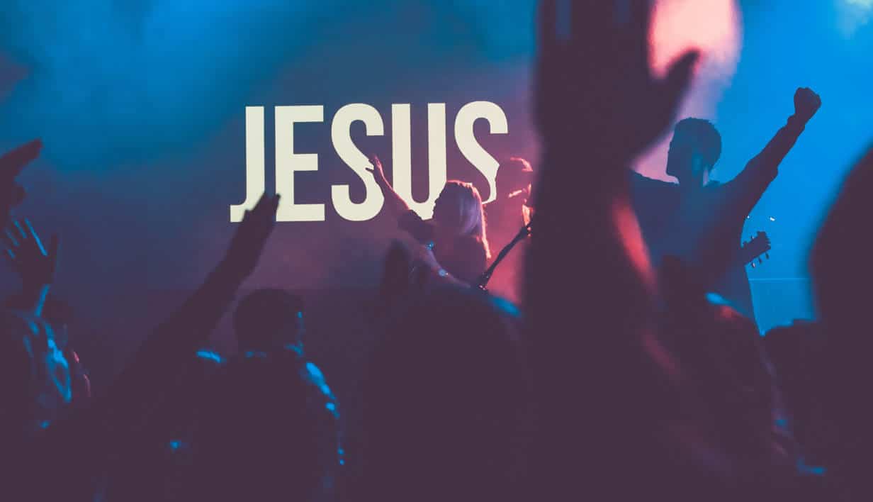 A crowd of worshippers looking at a stage with a singer holding up her hand. The background behind the singer reads, "Jesus."