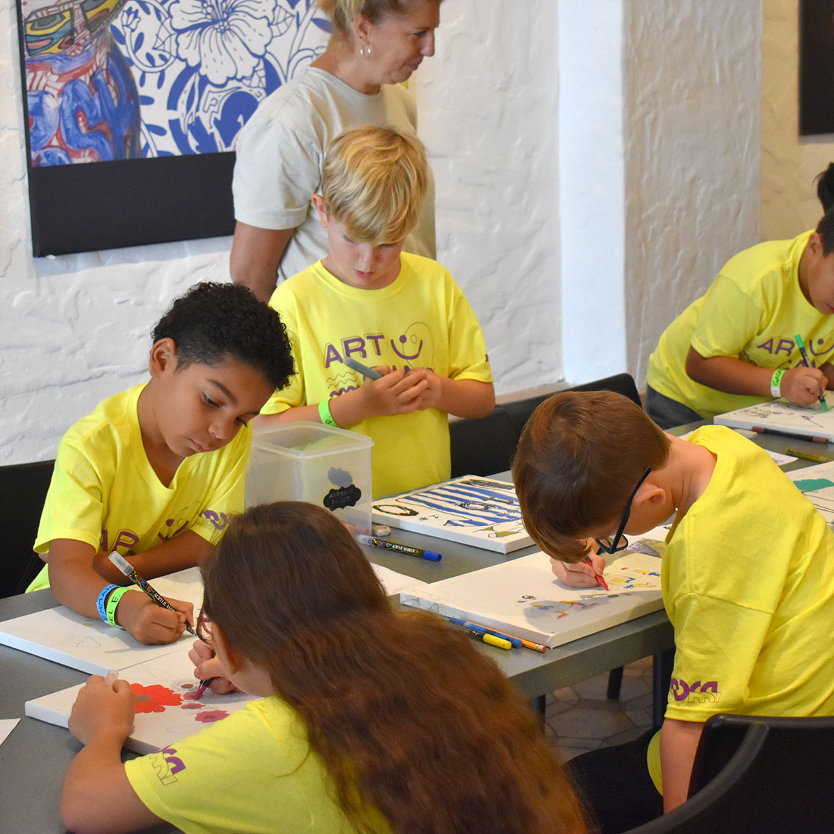 Un grupo de niños con camisetas amarillas brillantes del Campamento de Verano se sientan alrededor de una mesa y crean obras de arte con rotuladores.