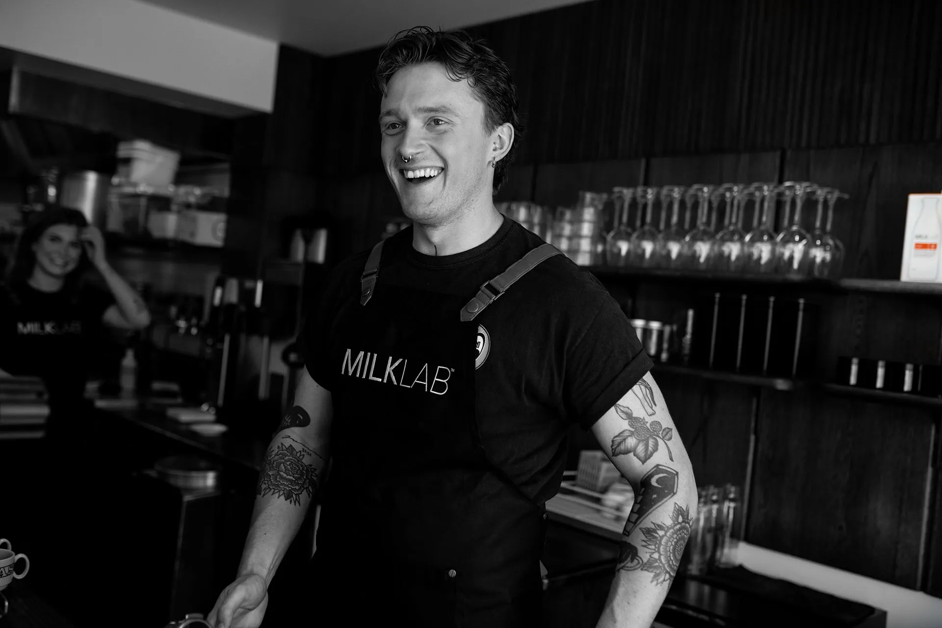 A male barista laughing wearing a MILKLAB apron with a second female barista in the background smiling