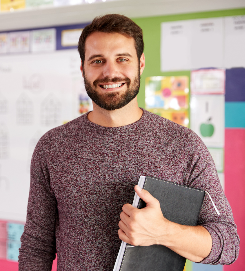 A smiling substitute teacher with a beard holding a leather-bound notebook in a classroom.