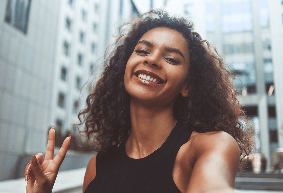 A Black woman with long hair is taking a selfie of herself while smiling and holding up a peace sign with her other hand. She's wearing a black tanktop. There are city buildings behind her.