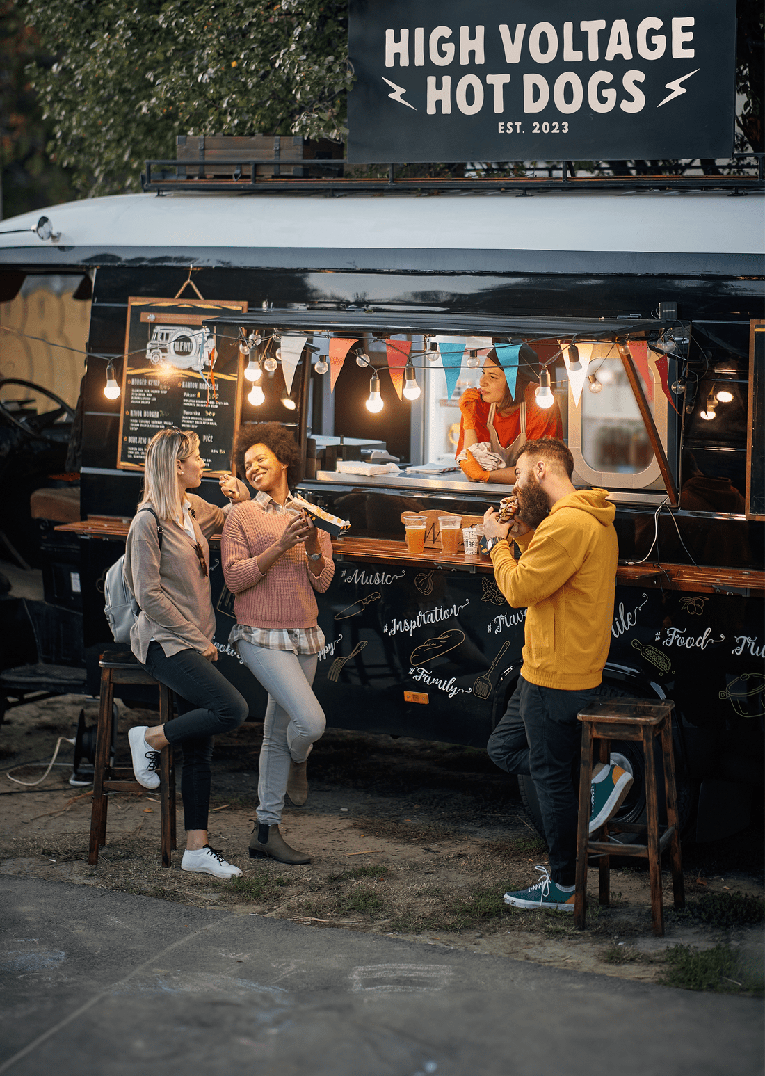 Three people laugh as they eat food next to a lively food truck with bistro lights.