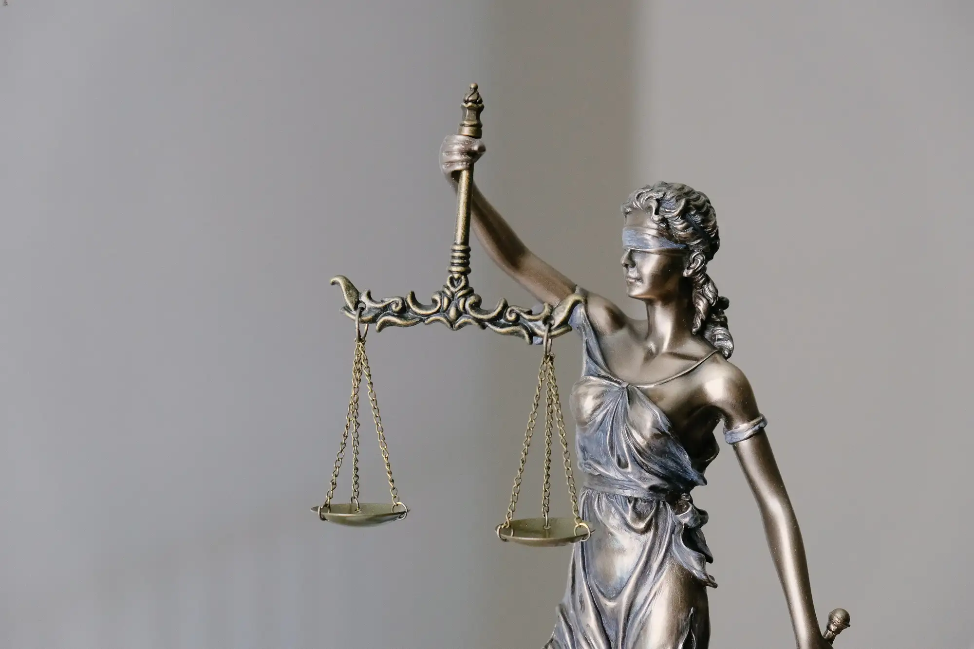 The image features a close-up of a statue of Lady Justice, a symbol of the judicial system, with a blindfold over her eyes, scales held high in her left hand, and a sword by her side. The statue is crafted in metallic tones, set against a soft-focus, neutral background that does not distract from the figure. The scales are balanced, representing the fair and equal administration of the law without bias or prejudice.
