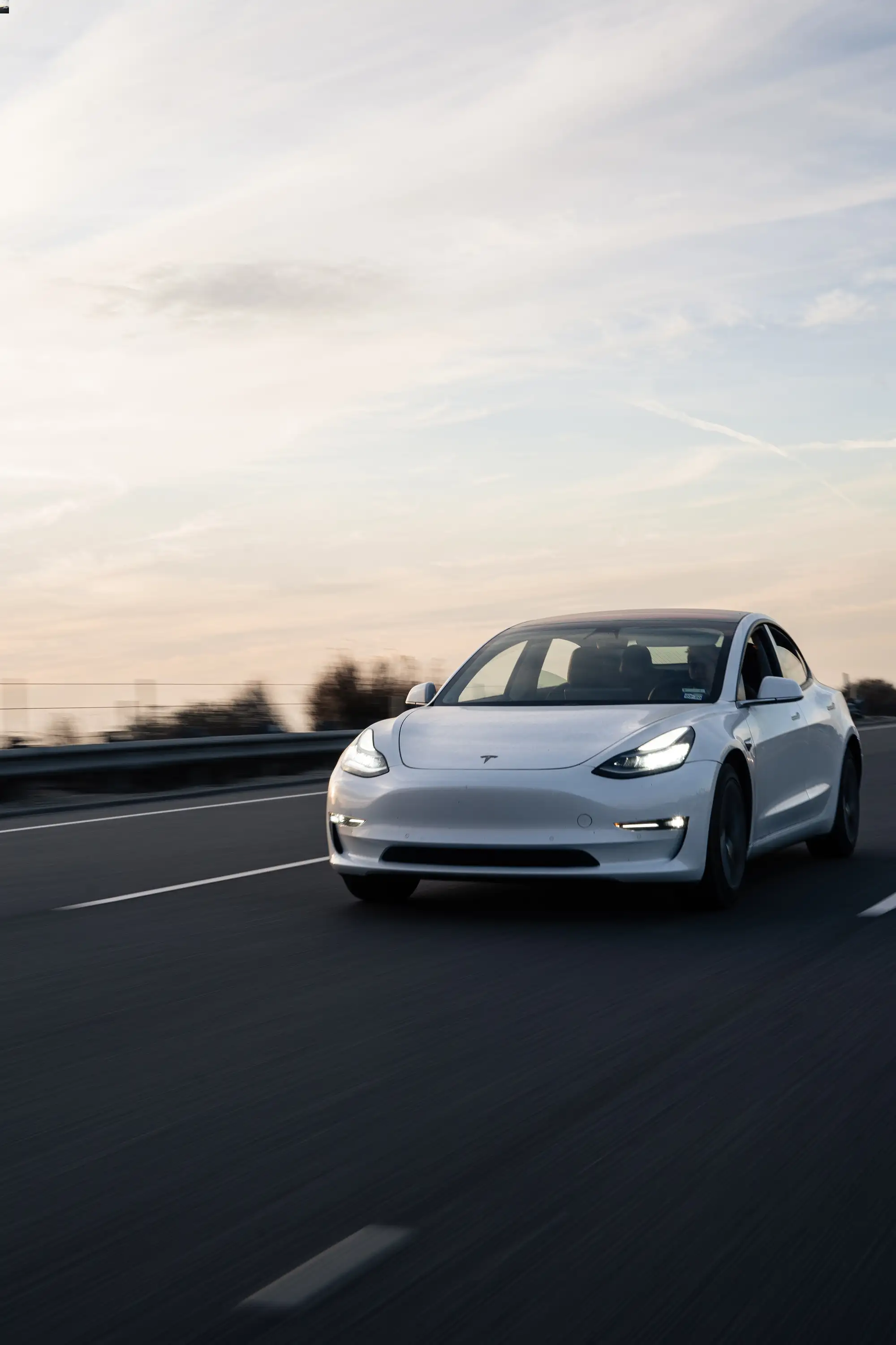 The image shows a white Tesla car in motion on a highway, captured with a sense of speed as the background blurs past. The sky above is a soft gradient of blues and whites, suggesting either dawn or dusk. The car