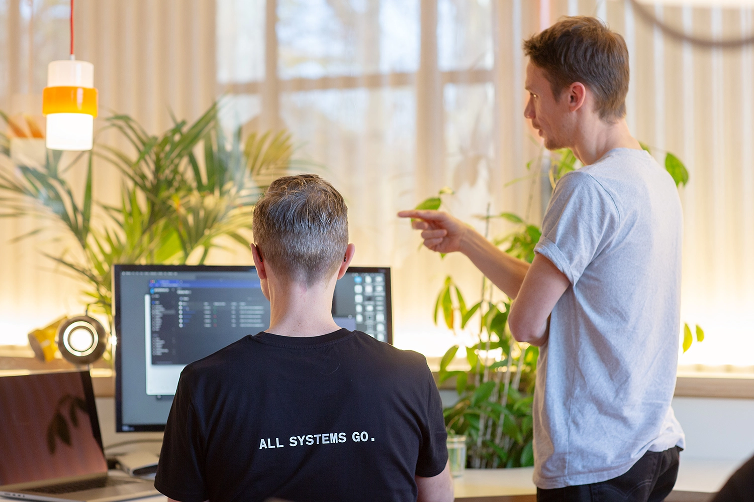 Daniel (Director) sat at his computer, with 'All Systems Go' on the back of his t-shirt. Charlie is stood next to him, pointing at his screen.