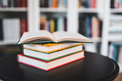 3 bunte geschlossene Bücher auf Tisch mit einem offenen Buch vor Bücherregal