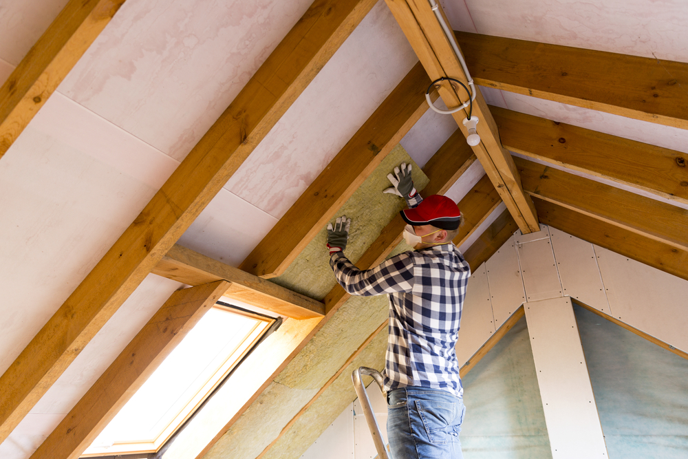 Dachdecker Handwerker steht auf Leiter unter Dachstuhl aus Holzbalken und platziert Glaswolle als Dämmungl