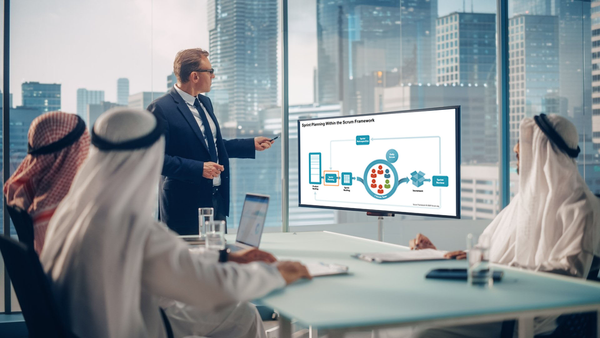 An office environment scene in the Middle East. There are views out of the window of office towers. In the foreground are four people dressed in business or Middle Eastern attire. One of the people is pointing to a TV screen as he presents - the topic on the screen is the Scrum Framework