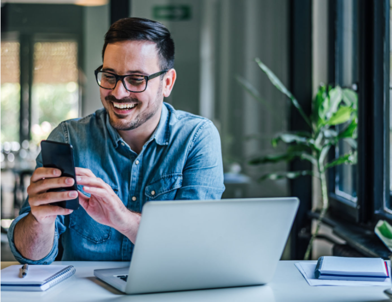 A man sat at his desk with his laptop open. He is smiling whilst he looks at his cell phone screen