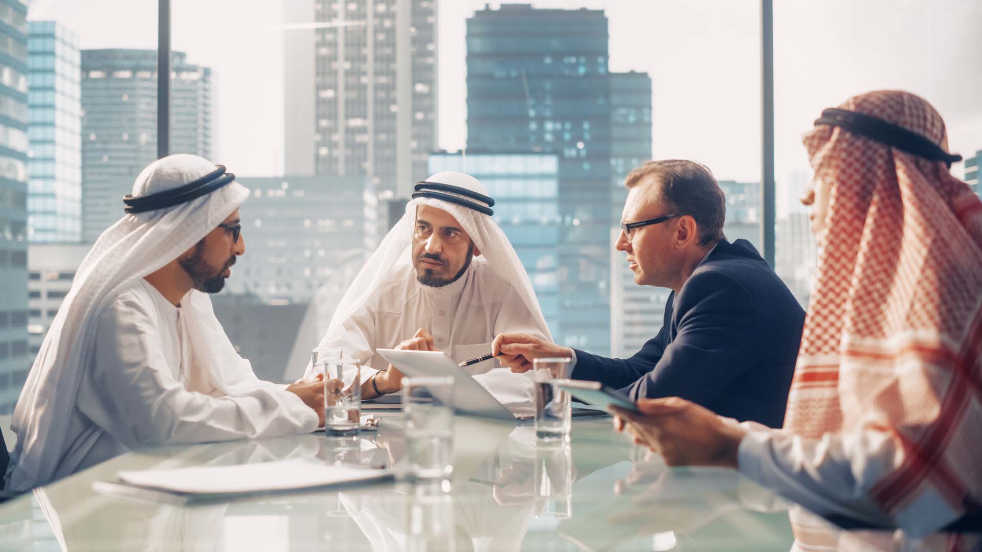 Four men sat around a desk, deep in discussion. A western looking man is pointing to a laptop screen with 3 other men around him, all dressed in Middle Eastern attire.