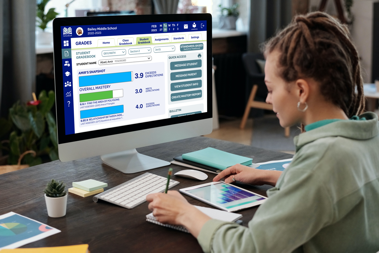 Person sitting in front of a desktop with a tablet and notebook in front of them; the desktop shows the Student Gradebook page