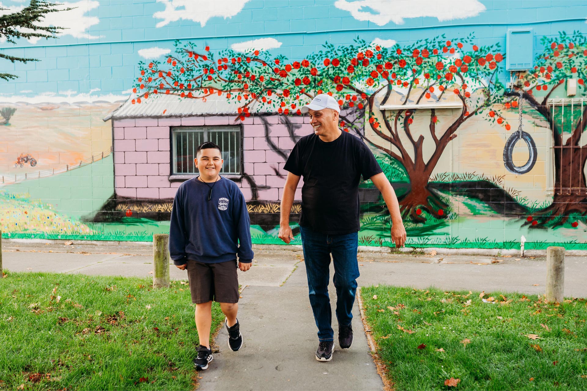 Man walking next to boy, both smiling with a colourful grafitti covered wall in the background.
