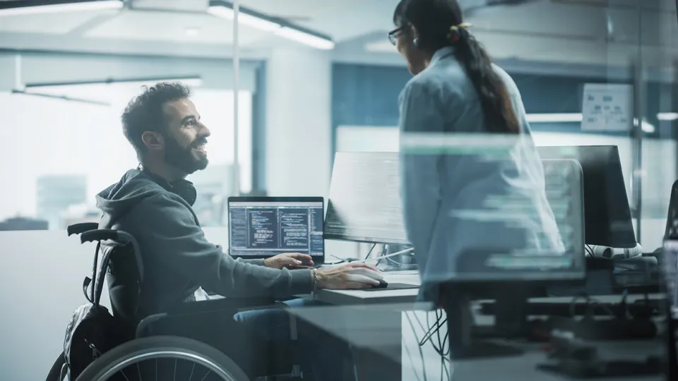 a man in a wheelchair working on a computer while talking to a woman
