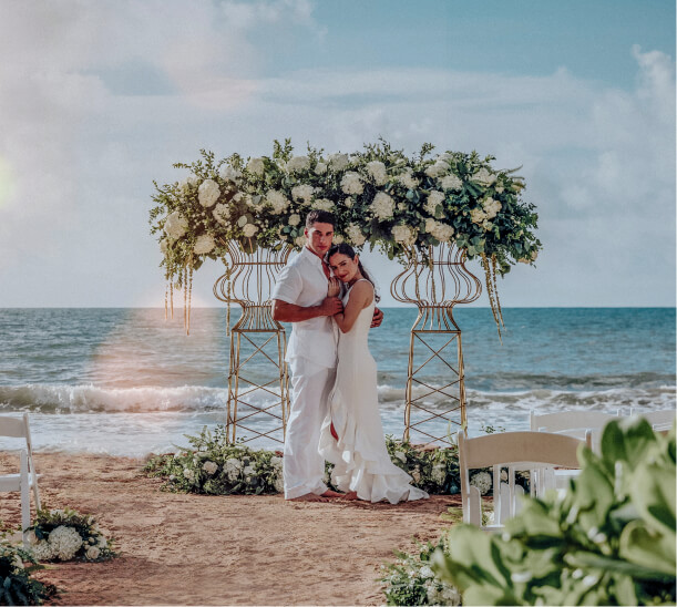 A couple dressed in all white posing in front of a wedding arch on the beach