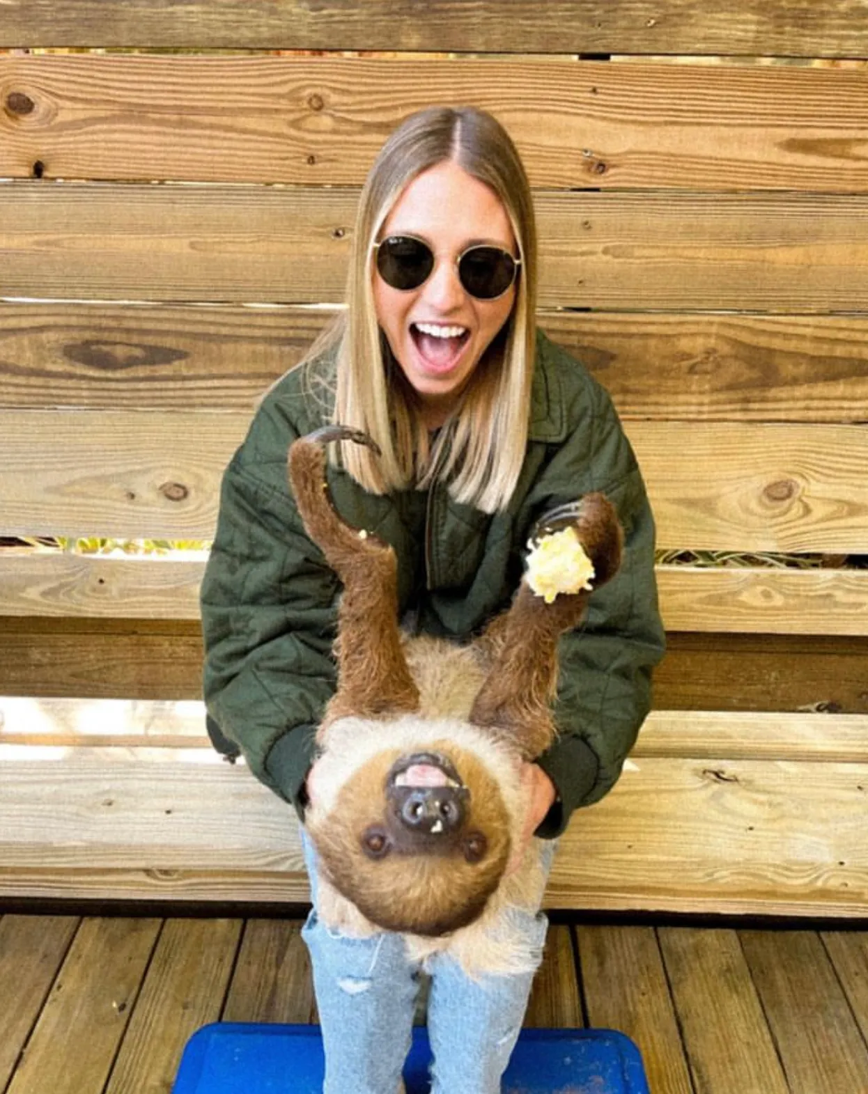 A woman wearing sunglasses holds a happy sloth in her lap