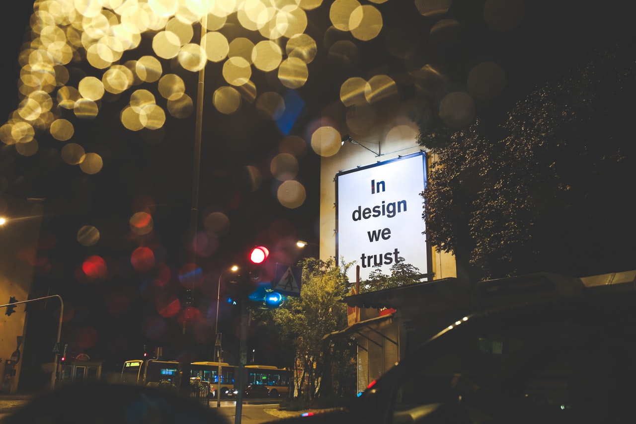 Nighttime city street photo of a billboard that reads "In Design We Trust"