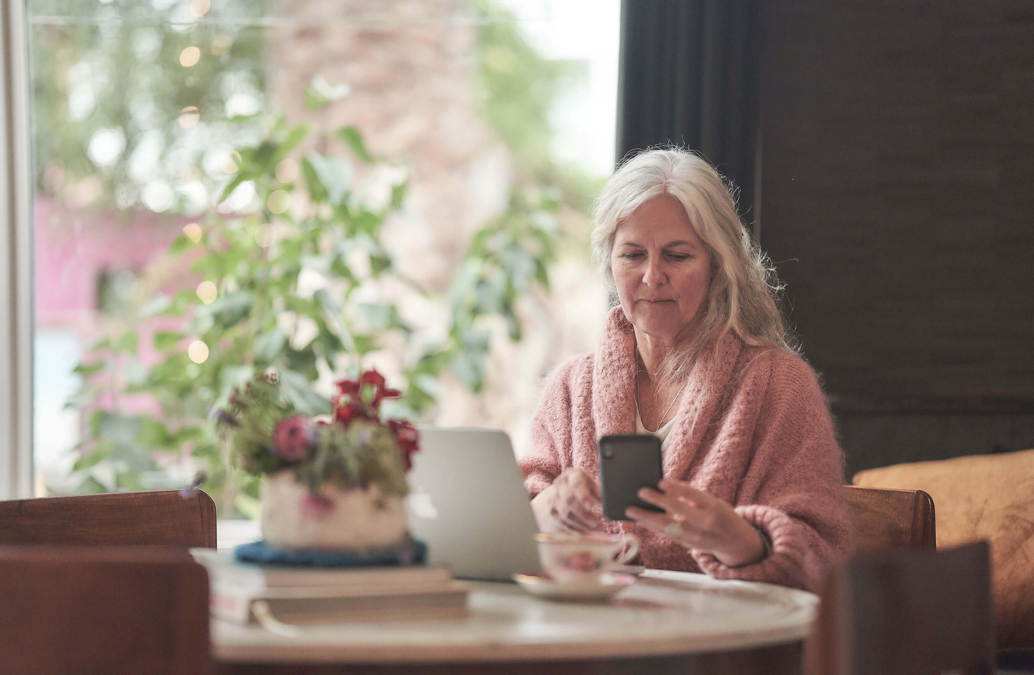 An elderly woman checks her phone at a table. Beside her, is a laptop, a pot with flowers and a teacup with a saucer