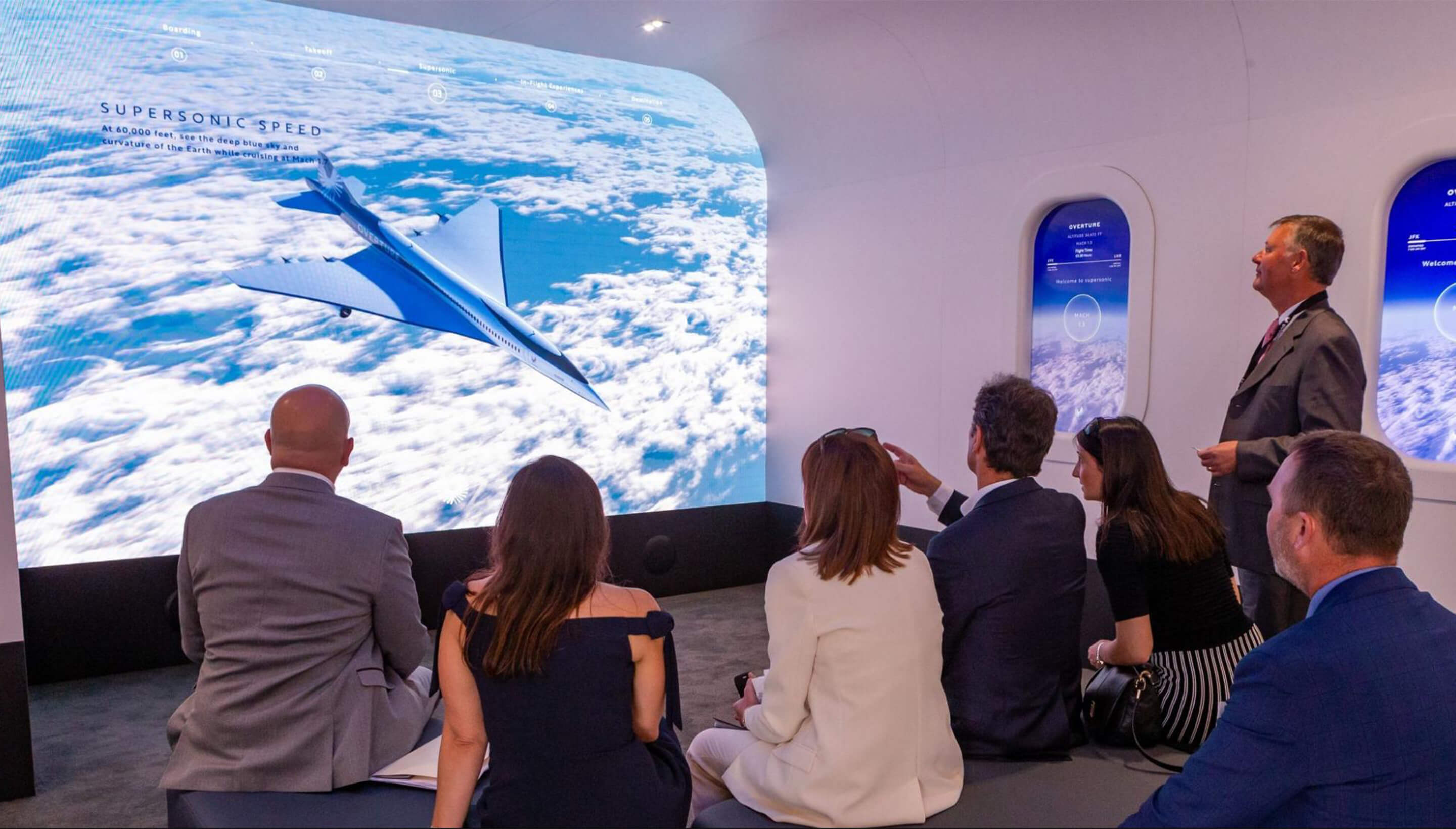 A group of people are in a room resembling an airplane, watching a supersonic jet being projected onto the wall