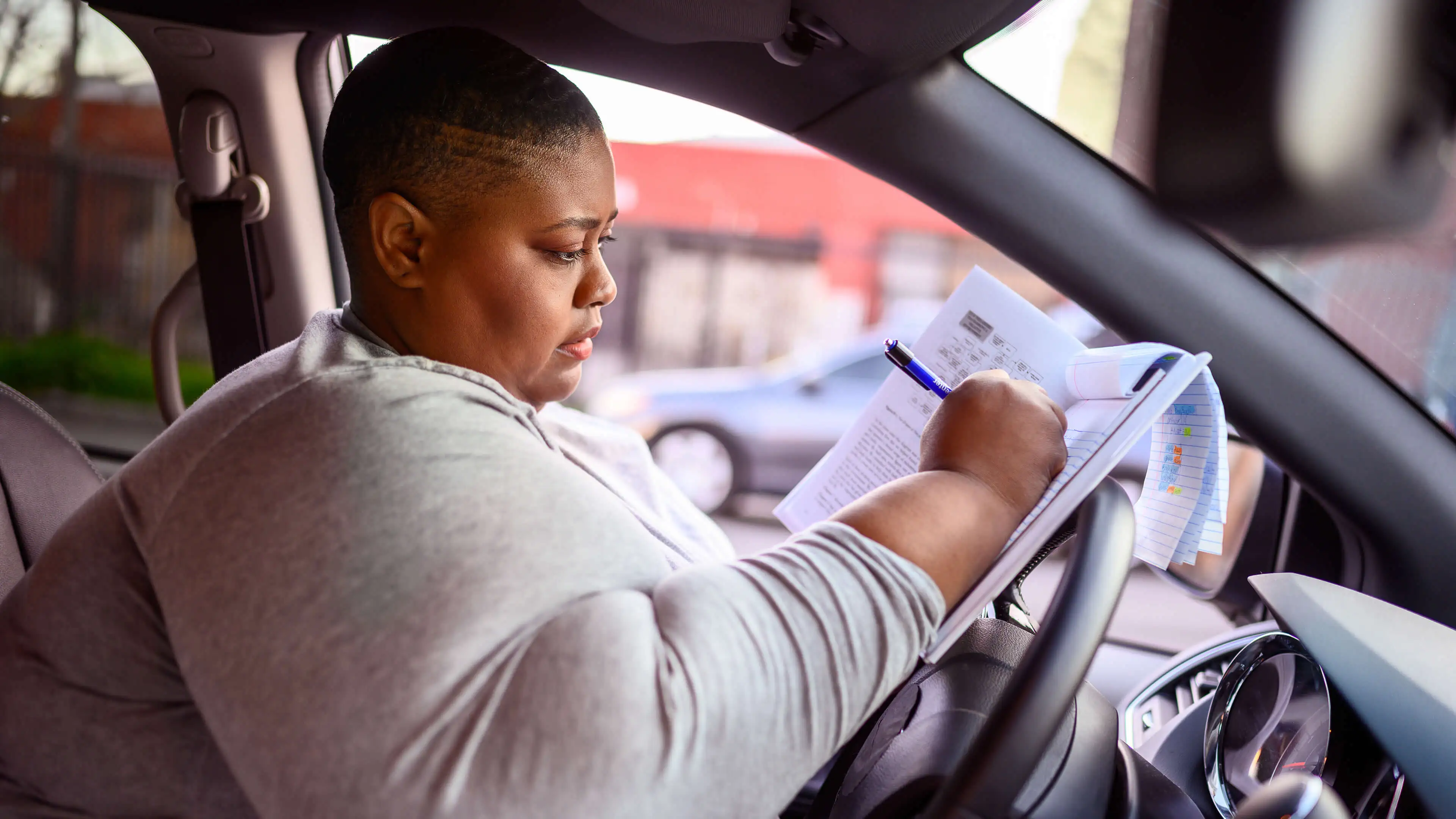 Black woman sitting in her car filling out paperwork on top of her steering wheel