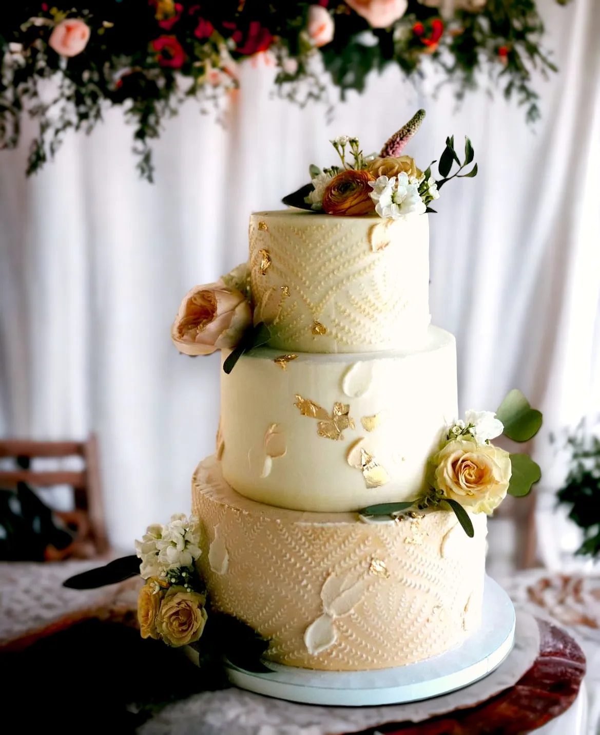 Three-tiered elegant cream and gold colored wedding cake with intricate pattern designs and yellow floral decorations.