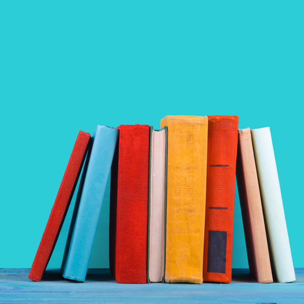 Eight multi-colored books leaning on each other on a blue shelf in front of a teal wall. 