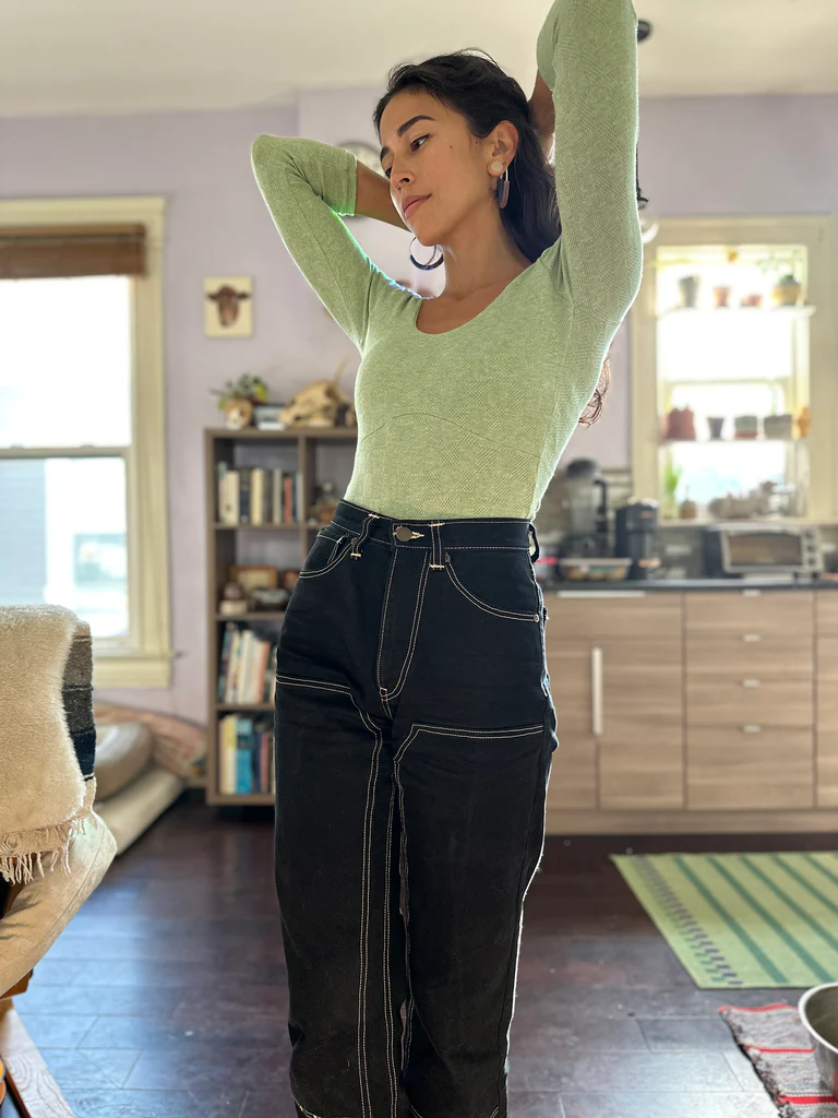 Leila, a small framed multiracial woman with dark wavy hair, stands in a living room. She is wearing a light green top and black jeans.