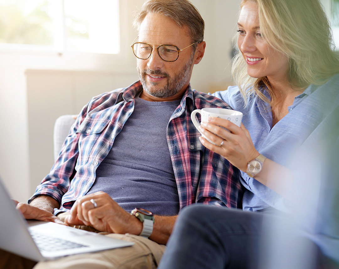 A middle-aged couple is in the living room, looking at the laptop. The woman is drinking coffee, and they are both looking at the screen very interested.