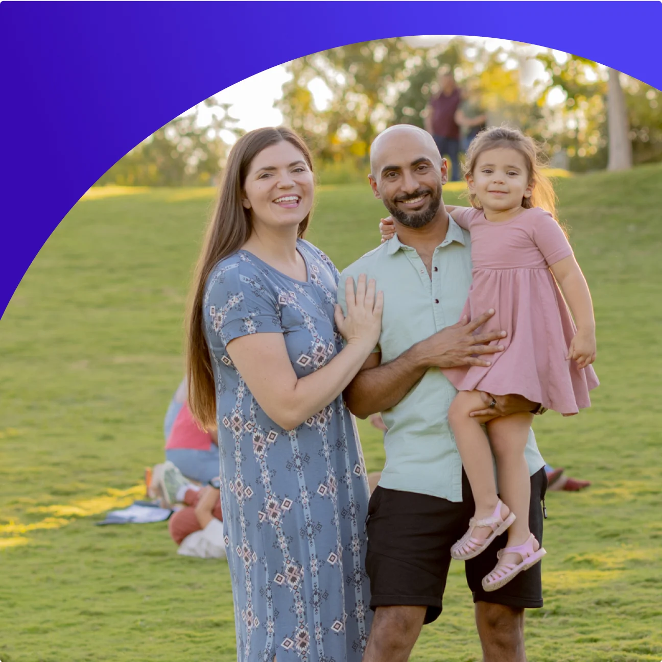 A couple holding a child in a pink dress on a green lawn. 