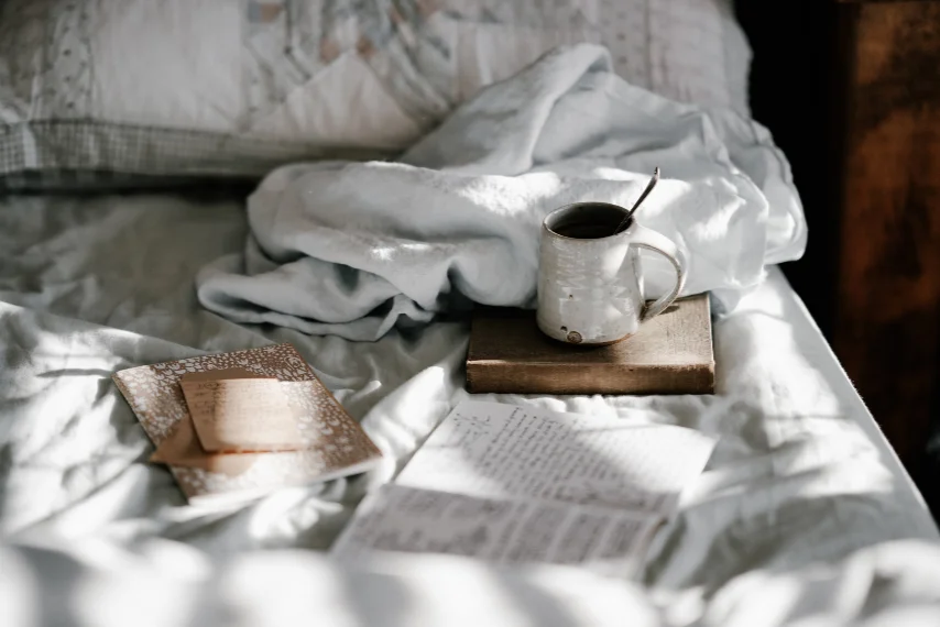 A cup of coffee on a book and papers on a bed.