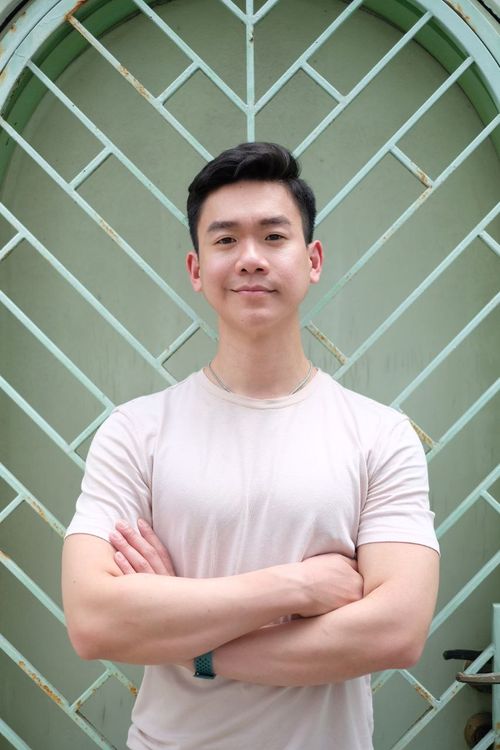 Asian actor Matheus Ting standing with his arms folded in front of an art deco gate.
