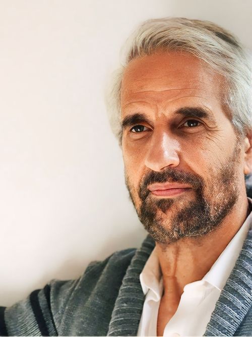 A portrait of a middle-aged man. He has grey hair and beard. He is dressed casually. He has quiet look
