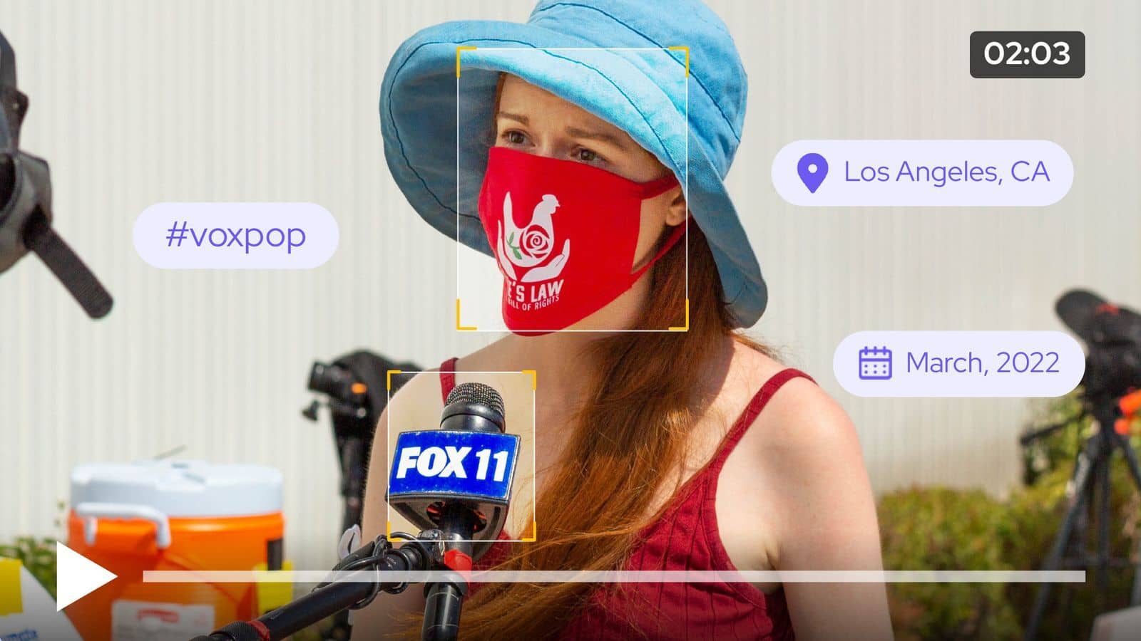 A video still image of a woman wearing a blue hat and red face mask being interviewed in the street, there is a microphone in front of her face and tags around her photo indicating 'vox pop', the location of LA, detection of a logo on the mic and her mask and the date of March 2022 is depicted..