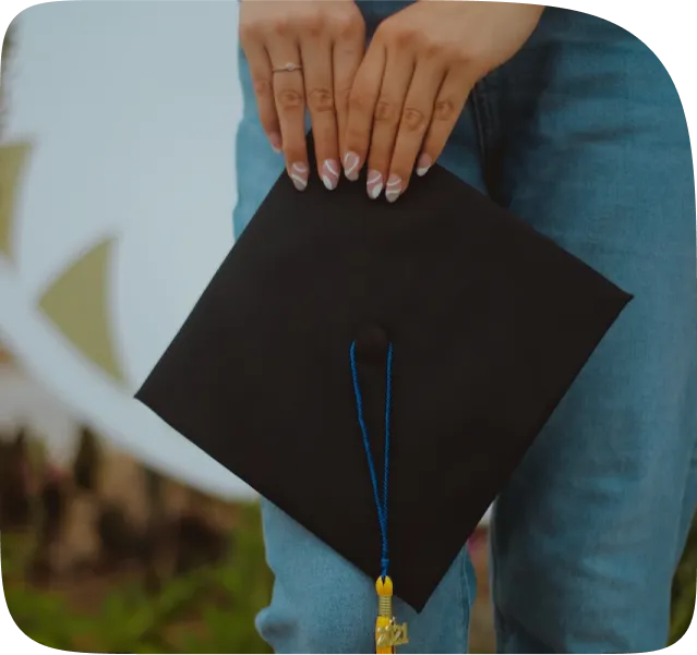 A shot of a woman's hands holding a graduation cap. She's standing up, wearing jeans. 